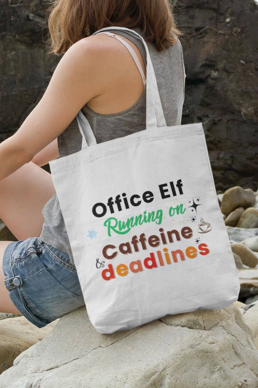 Office tote bag with a person holding a tote bag with humorous text 'Office Elf, Running on Caffeine & Deadlines' on a rocky surface
