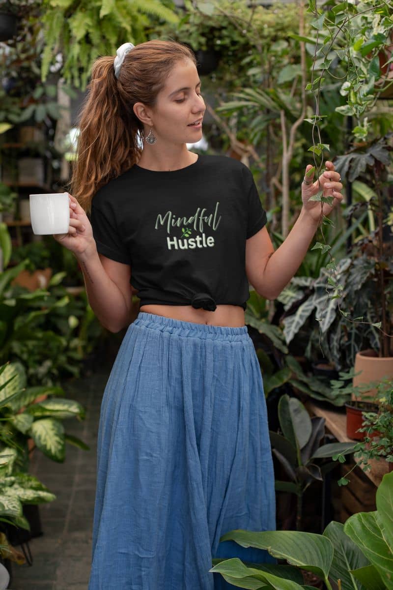 Positive quote tee with a woman holding a mug and a plant in a garden setting, wearing a black t-shirt with 'Mindful Hustle' text.