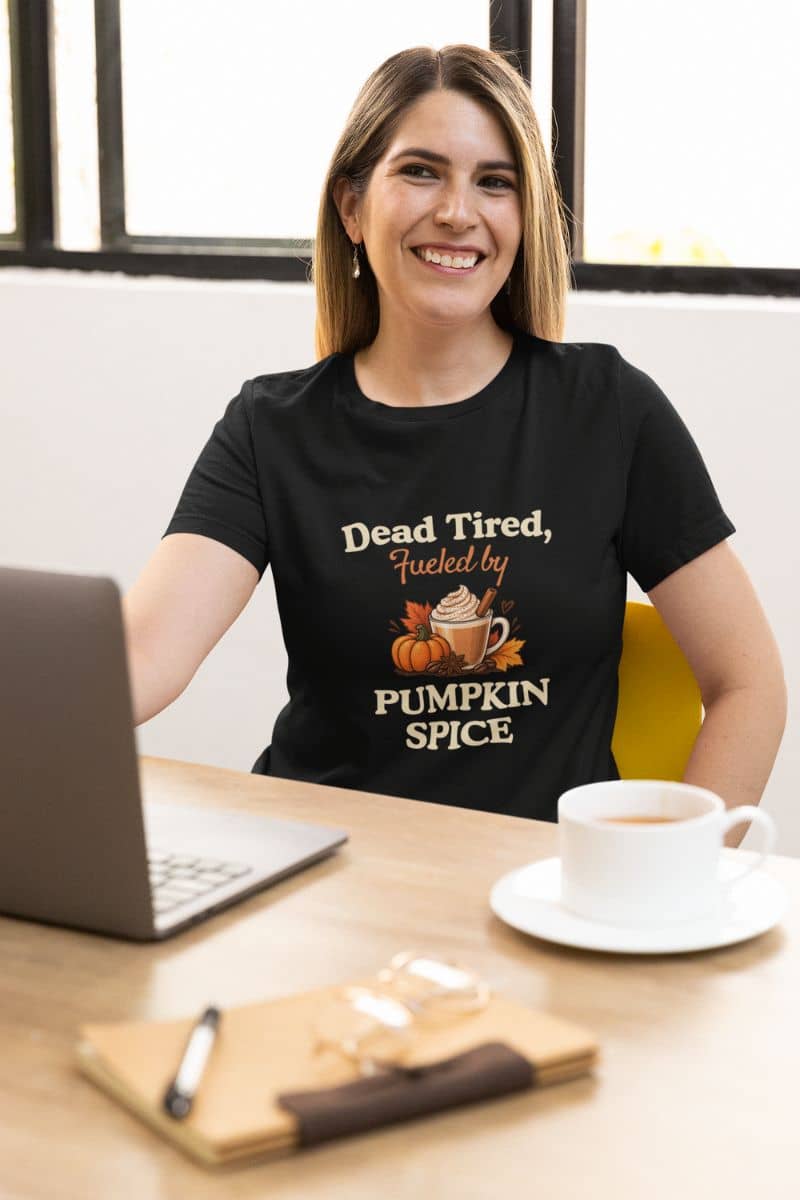 Pumpkin spice season t shirt with a smiling woman sits at a desk wearing a black graphic T-shirt that reads 'Dead Tired, Fueled by Pumpkin Spice' with a small autumn illustration. 