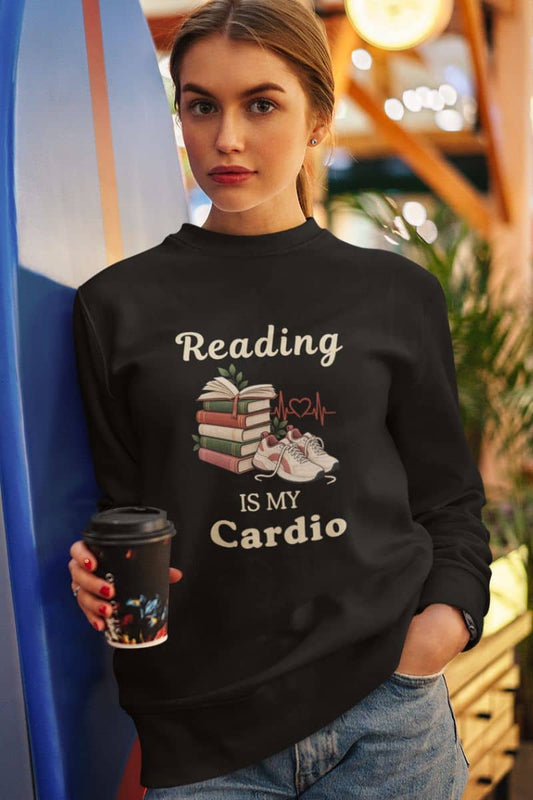Reading sweatshirt with a woman wearing a black sweatshirt that reads 'Reading Is My Cardio'. She is holding a coffee cup and standing in a warm, bokeh-lit indoor environment.