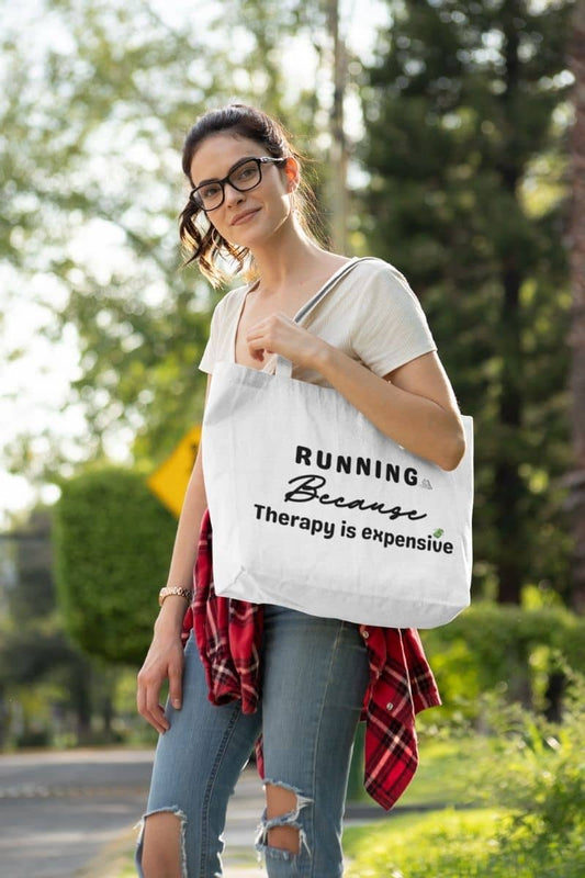 Running quote tote bag with a woman holding a tote with text 'Running because therapy is expensive', outdoors