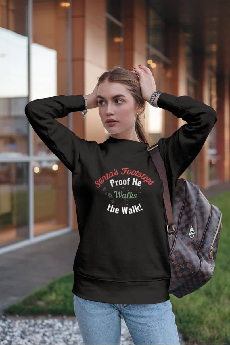 Santa footprint sweatshirt with a woman wearing a black sweatshirt with text 'Santa's Footsteps - Proof He Walks The Walk!', standing outdoors.