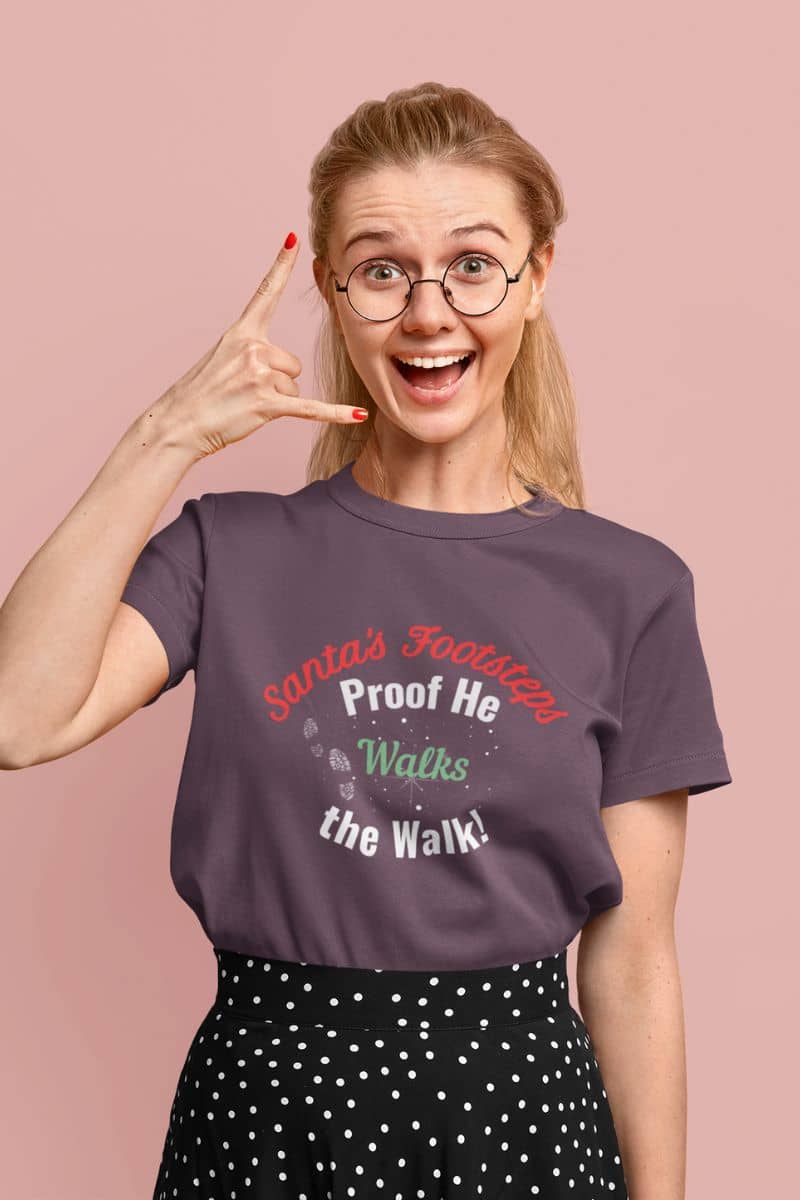 Santa proof shirt with a woman wearing a purple t-shirt with text 'Santa's Footsteps - Proof He Walks The Walk!', making a peace sign against a pink background