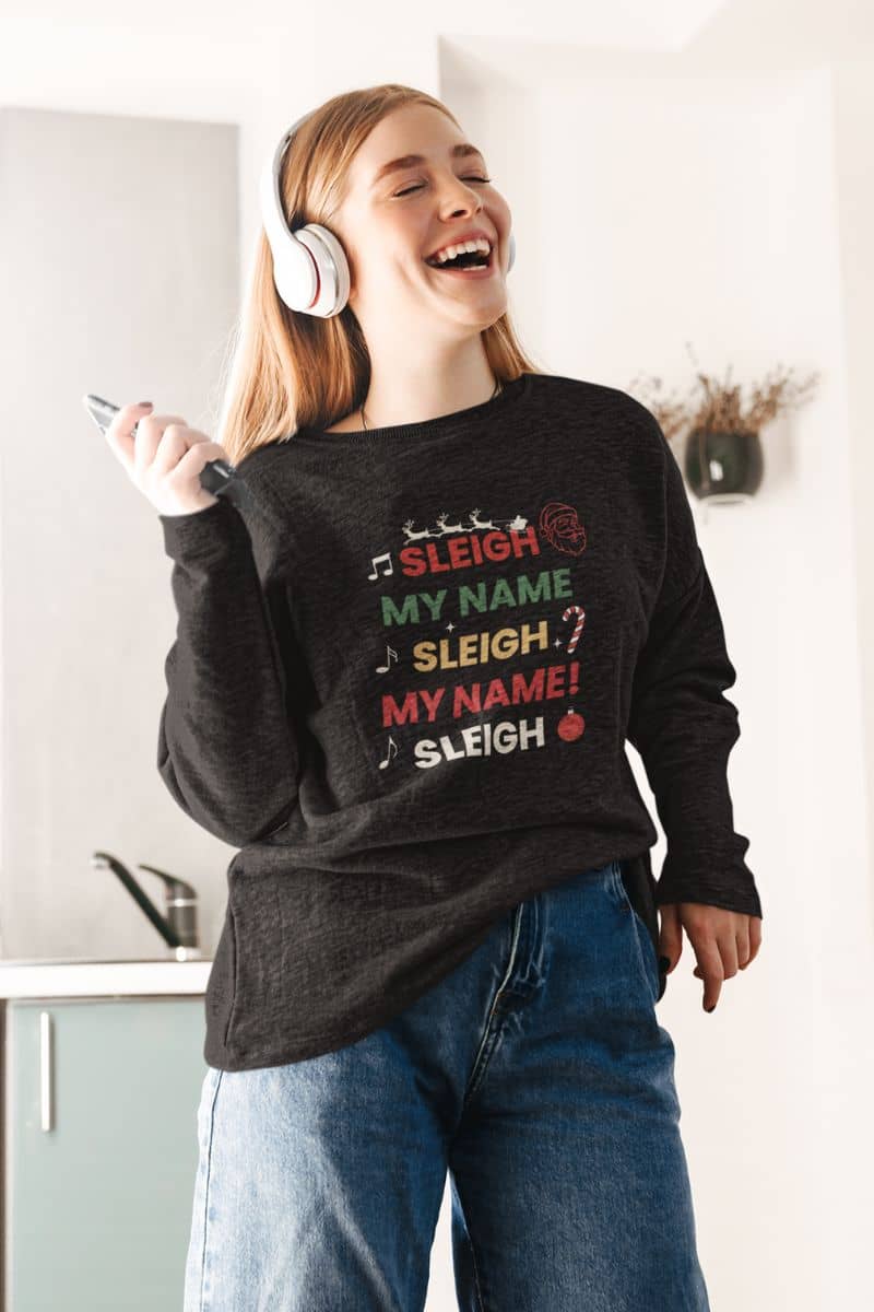 Santa sleigh sweater with a person wearing a black sweatshirt with text 'Sleigh My Name, Sleigh My Name' and headphones, standing in a kitchen.