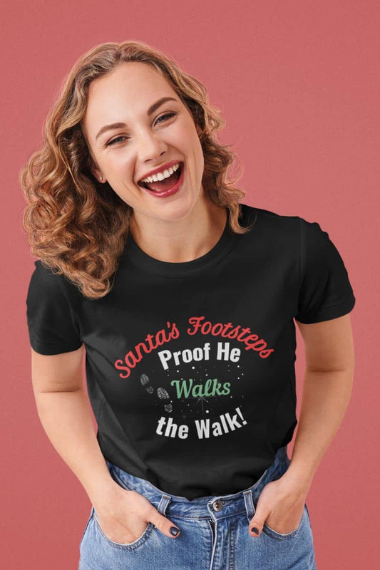 Santa's footsteps t-shirt with a woman wearing a black t-shirt with humorous text 'Santa's Footsteps - Proof He Walks The Walk!' on a pink background