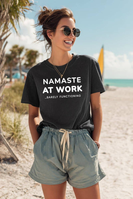 Sarcastic office shirt with a woman wearing a black t-shirt with text 'Namaste at Work Barely Functioning' on a beach.
