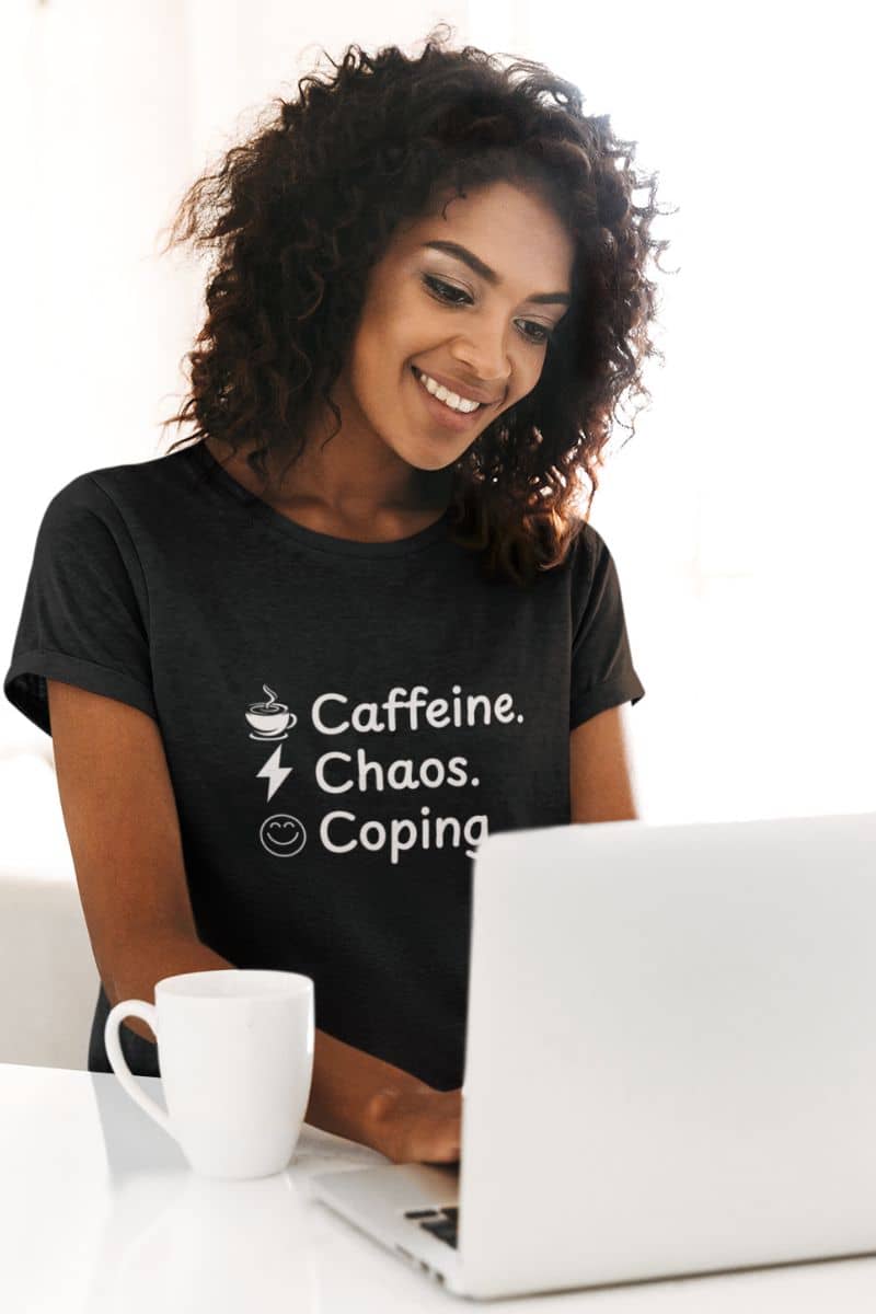 Sarcastic quote t-shirt with a woman wearing a black t-shirt with text 'Caffeine, Chaos & Coping', using a laptop at a desk.
