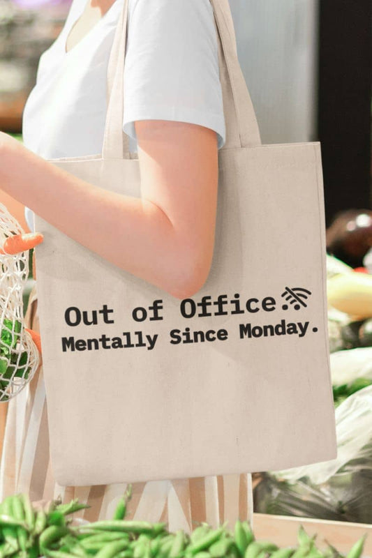 Sarcastic work tote bag featuring the text 'Out of Office. Mentally Since Monday,' held by a person in an outdoor setting.