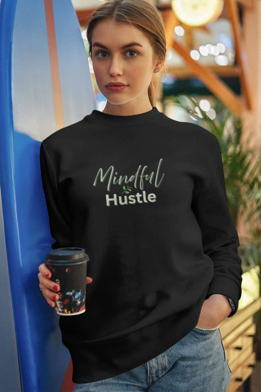 Self-development sweatshirt with a woman wearing a black sweatshirt with 'Mindful Hustle' text, holding a coffee cup, standing next to a blue surfboard.