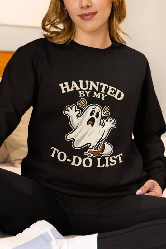 A woman wearing a spooky sweatshirt featuring a “Haunted by My To-Do List” funny ghost, sitting on the couch