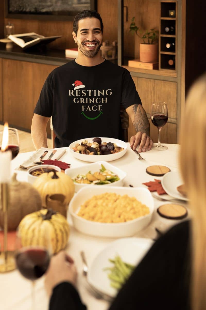 Subtle Christmas humor shirt with a man wearing a 'Resting Grinch Face' shirt at a holiday dinner table with food and wine.