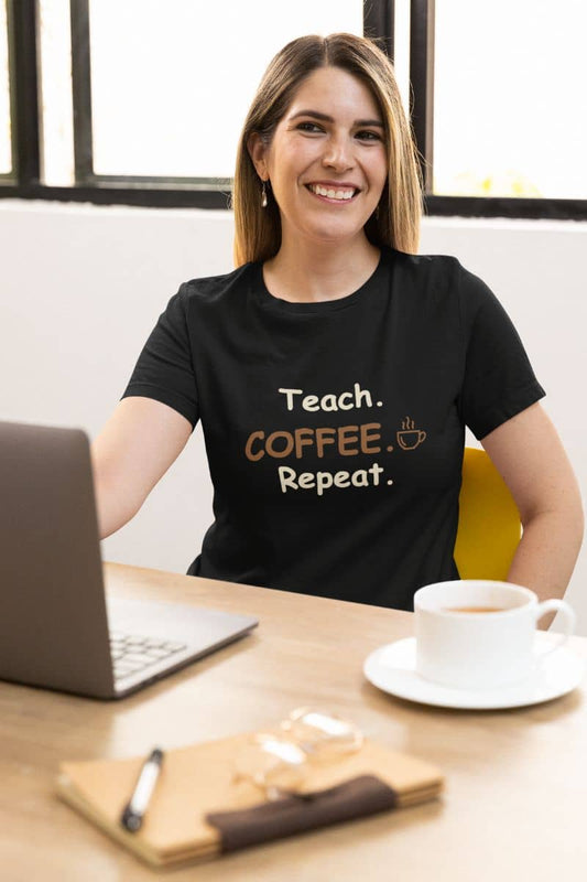 Teacher graphic tee with a smiling woman sits at a desk wearing a black graphic T-shirt that reads 'Teach. COFFEE Repeat.