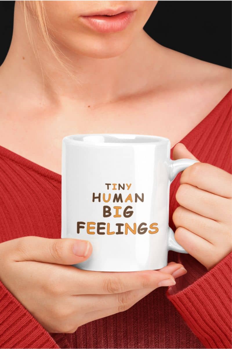 Teacher mug with a person in a red ribbed sweater holds a white ceramic mug with text 'Tiny Human Big Feelings' 