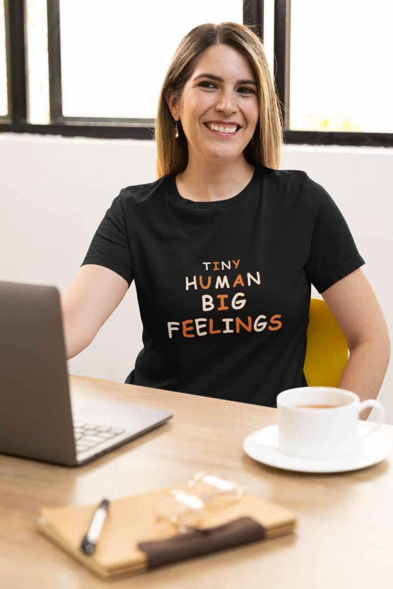 Teacher shirt with a woman smiling at a desk wearing a black graphic t-shirt that reads 'Tiny Human Big Feelings' in white and orange text. The scene includes a laptop, a coffee cup, and a notebook on a wooden desk.