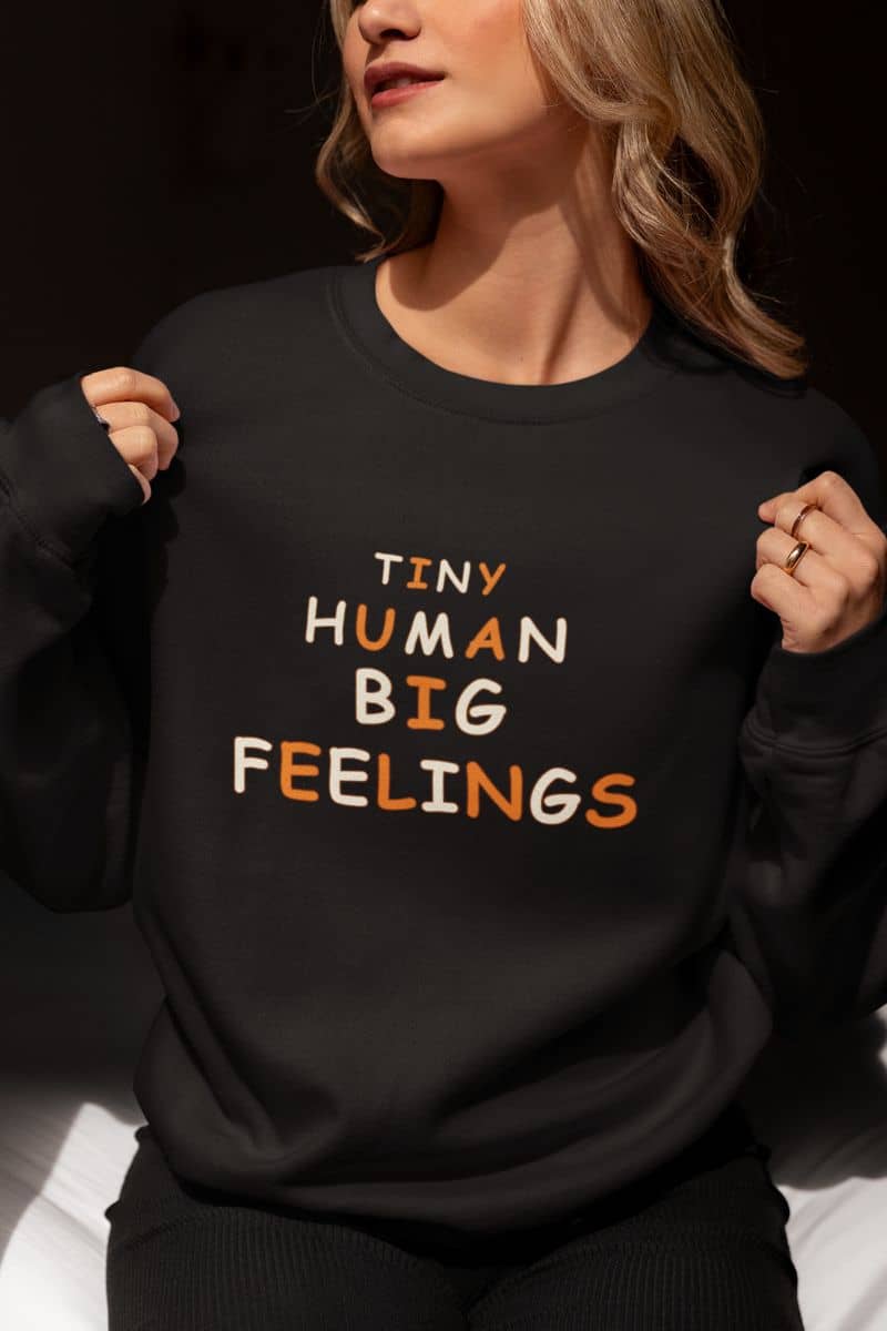 Teacher sweater with a woman wearing a black crewneck sweatshirt with text 'Tiny Human Big Feelings' 
