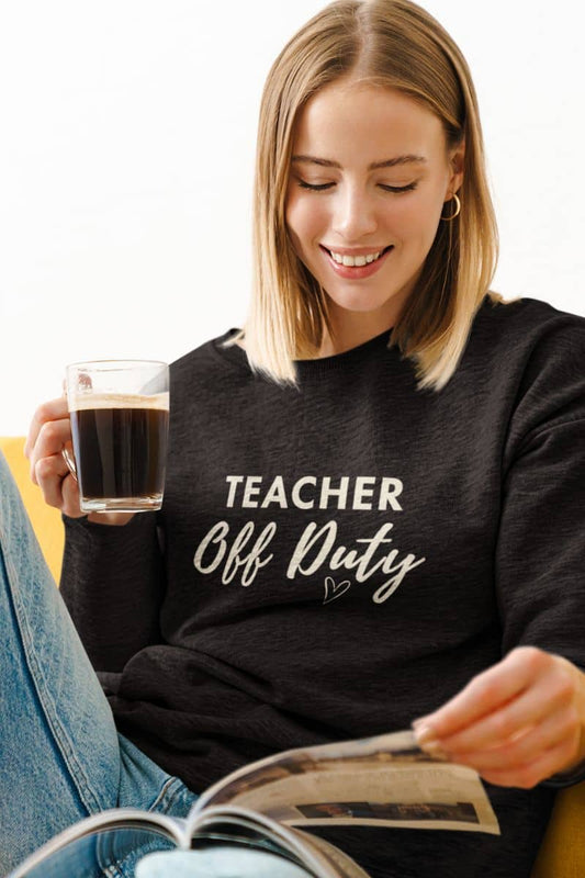 Teacher sweatshirt for women with a woman with blonde hair wearing a black crewneck sweatshirt featuring the text 'TEACHER Off Duty' printed in white on the front. She is holding a glass of coffee and looking at a magazine while sitting on a yellow chair.