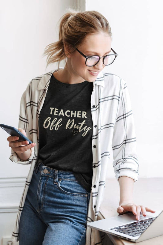 Teacher t shirt with a woman wearing a black t-shirt that reads 'TEACHER Off Duty', styled with a white and gray plaid flannel shirt and blue jeans. She is sitting at a desk using a laptop and holding a smartphone.