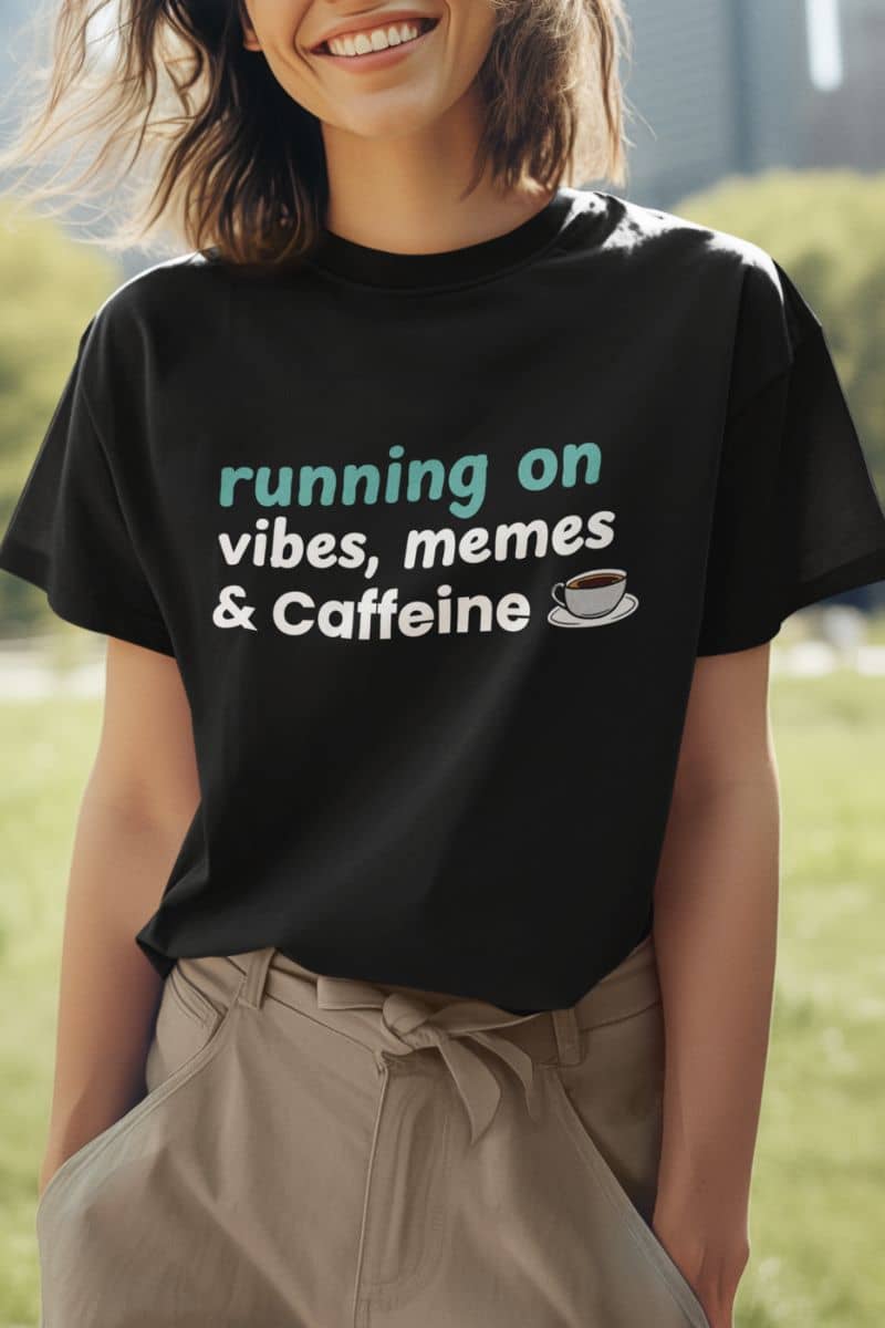 Tired but caffeinated shirt with a person wearing a black t-shirt with text 'running on vibes, memes & caffeine' in an outdoor setting.