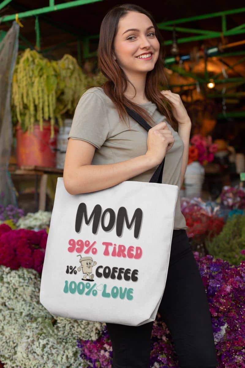 Tired mom tote bag with a woman holding a tote bag with text 'Mom 99% Tired, 1% Coffee, 100% Love' at an outdoor market.