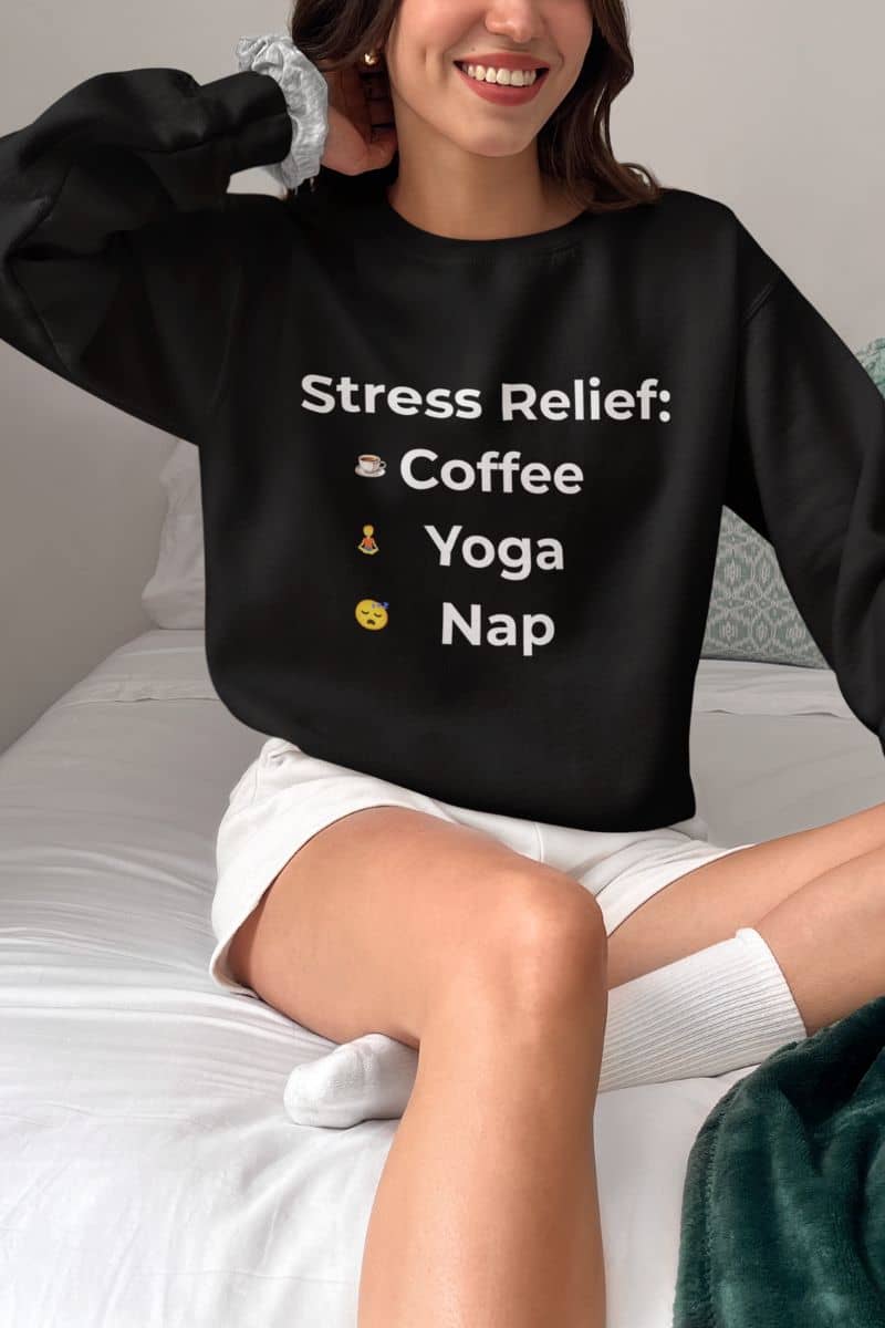 Trendy sweatshirt with a person wearing a black sweatshirt with 'Stress Relief: Coffee Yoga Nap' text, sitting on a bed.