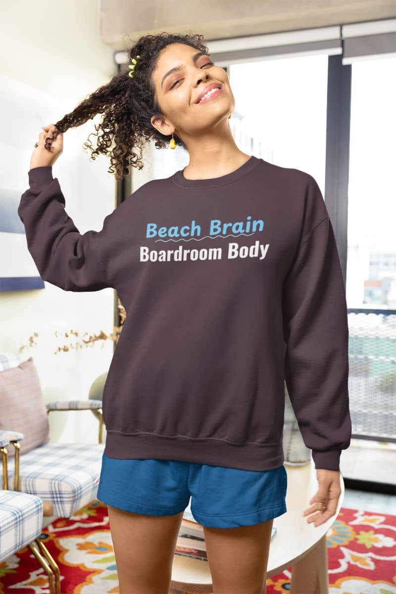 Vacation mood sweatshirt with a woman wearing a sweatshirt with 'Beach Brain Boardroom Body' text in a living room.