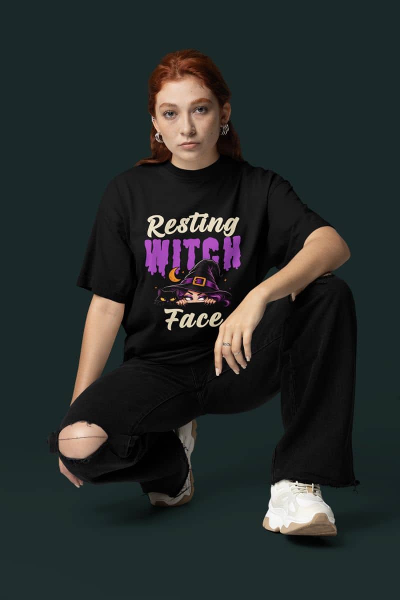Witchy graphic t shirt with a woman with red hair crouches against a dark green background wearing a black graphic t-shirt that reads 'Resting Witch Face' in purple and cream lettering, paired with black ripped jeans and white sneakers.