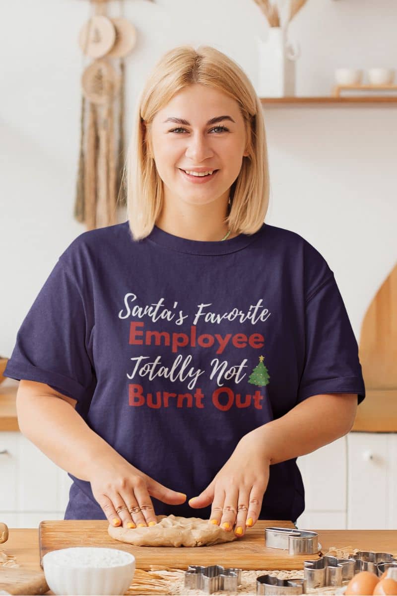 Work from home humor tee with a woman wearing a purple t-shirt with text 'Santa’s Favorite Employee Totally Not Burnt Out', in a kitchen
