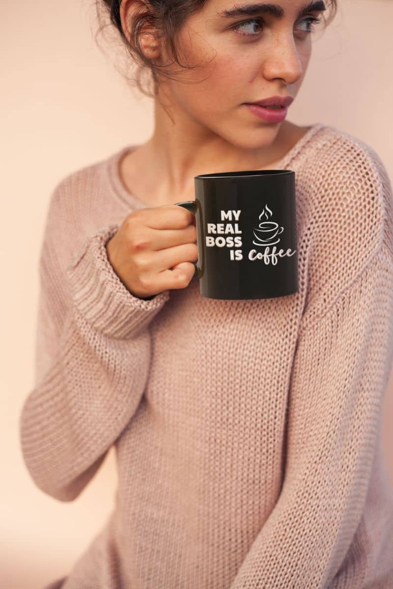 Work humor mug with a woman holding a black mug with 'My real boss is coffee' text against a plain background