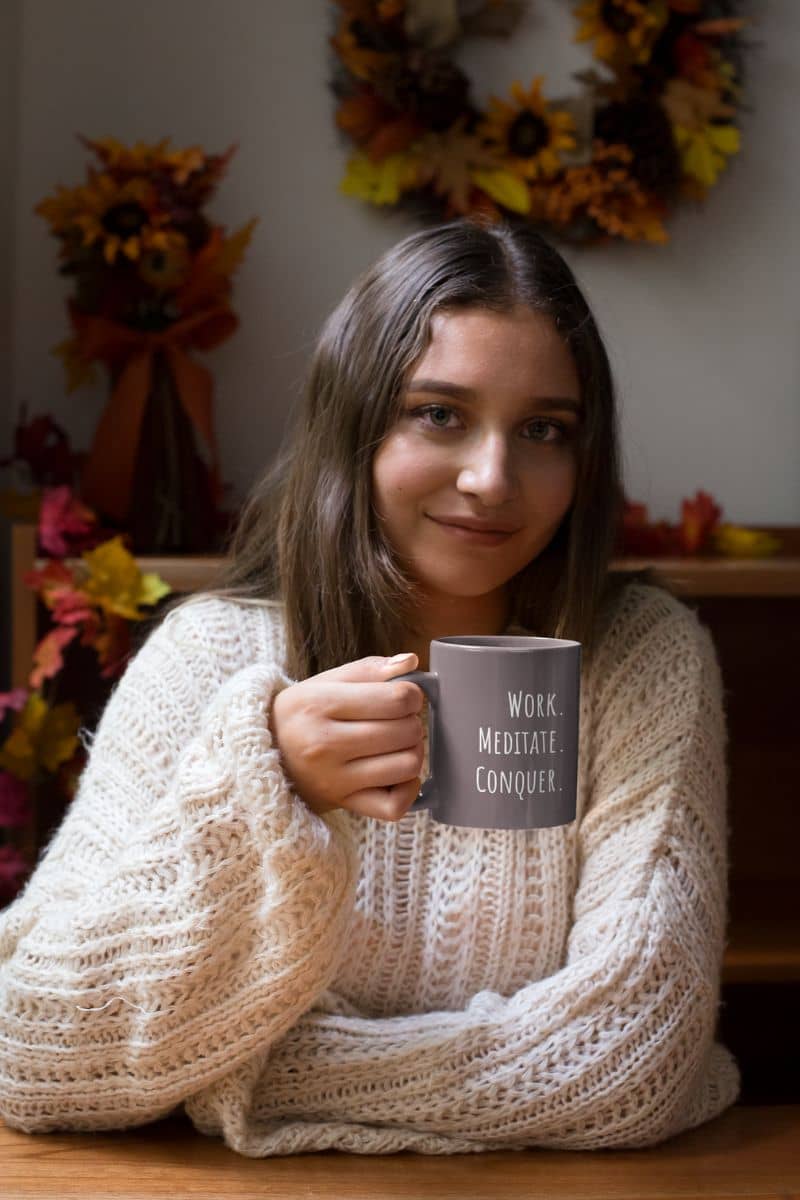 Work meditate conquer coffee mug with a young girl sitting at a table holding a cup of coffee in her hand