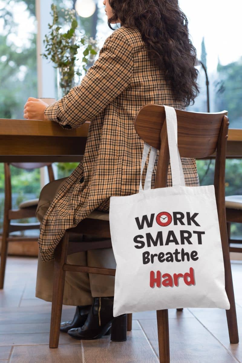 Work motivation tote bag with a person sitting at a table with a tote bag that says 'Work Smart Breathe Hard' on a blurred background.