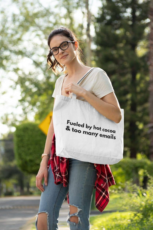 Workday humor tote bag with a woman holding a tote bag with text 'Fueled by hot cocoa & too many emails' outdoors