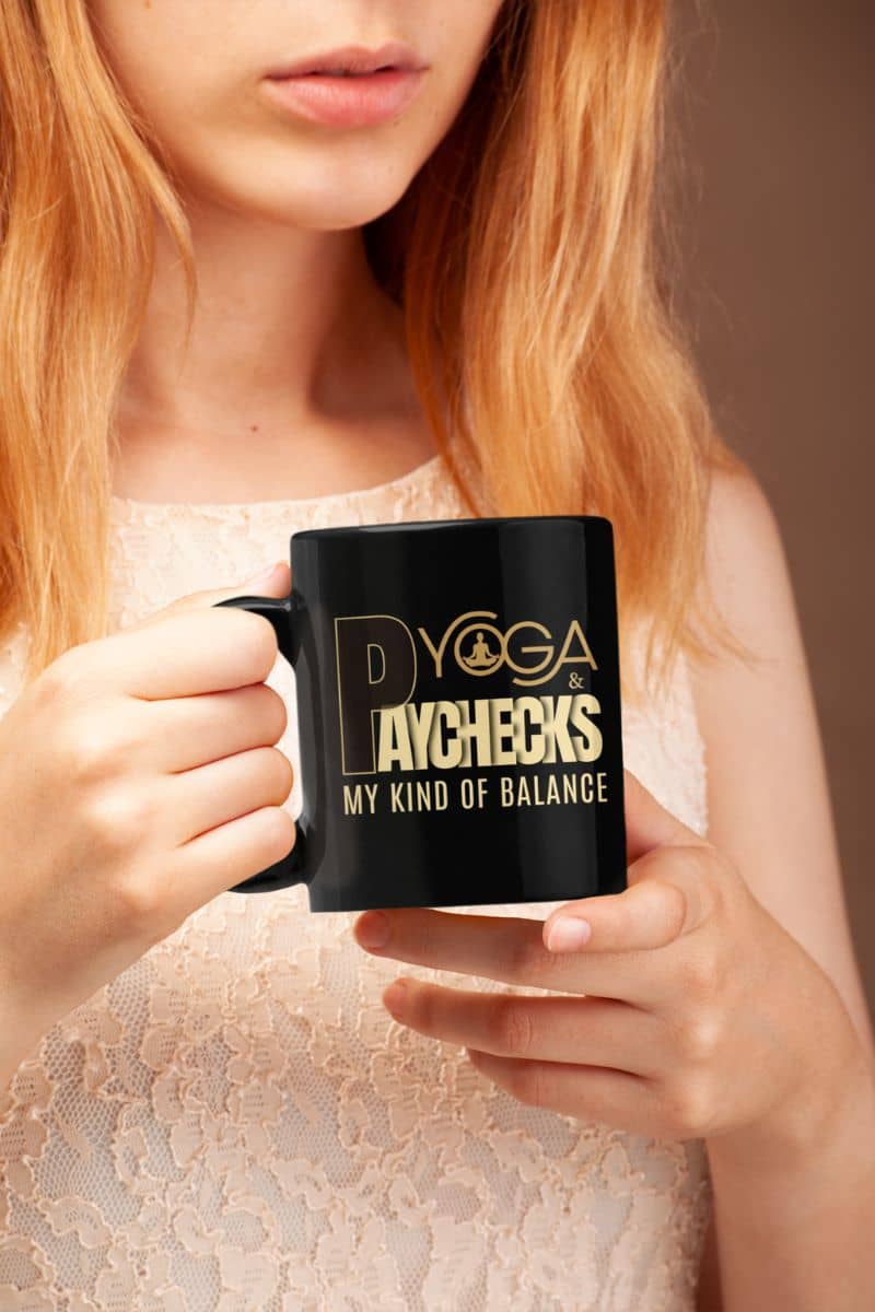 Yoga coffee mug for professionals with a person holding a black mug with gold text 'Yoga & Paychecks – My Kind of Balance', wearing a white lace top.
