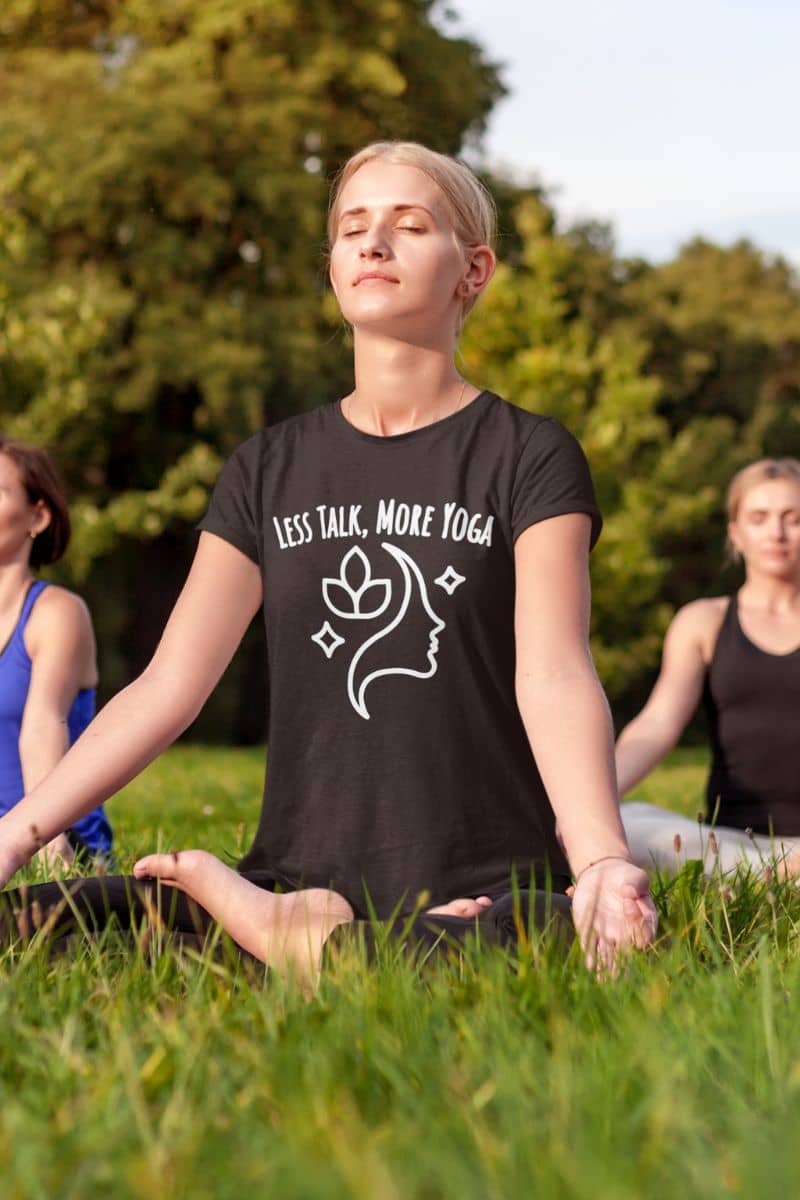 Yoga quote t-shirt with a person wearing a black t-shirt with text 'Less Talk, More Yoga' sitting on grass outdoors.