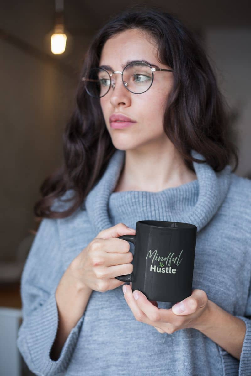 Zen morning mug with a woman holding a black mug with 'Meanful Hustle' text, wearing glasses and a gray sweater.