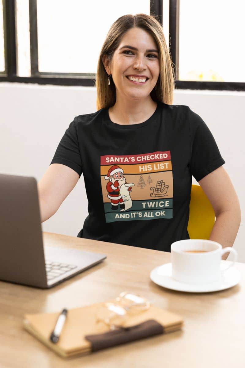 adult holiday t-shirt with a woman wearing a black t-shirt with text 'Santa's Checked His List Twice And It's All Ok', sitting at a desk with a laptop and coffee.