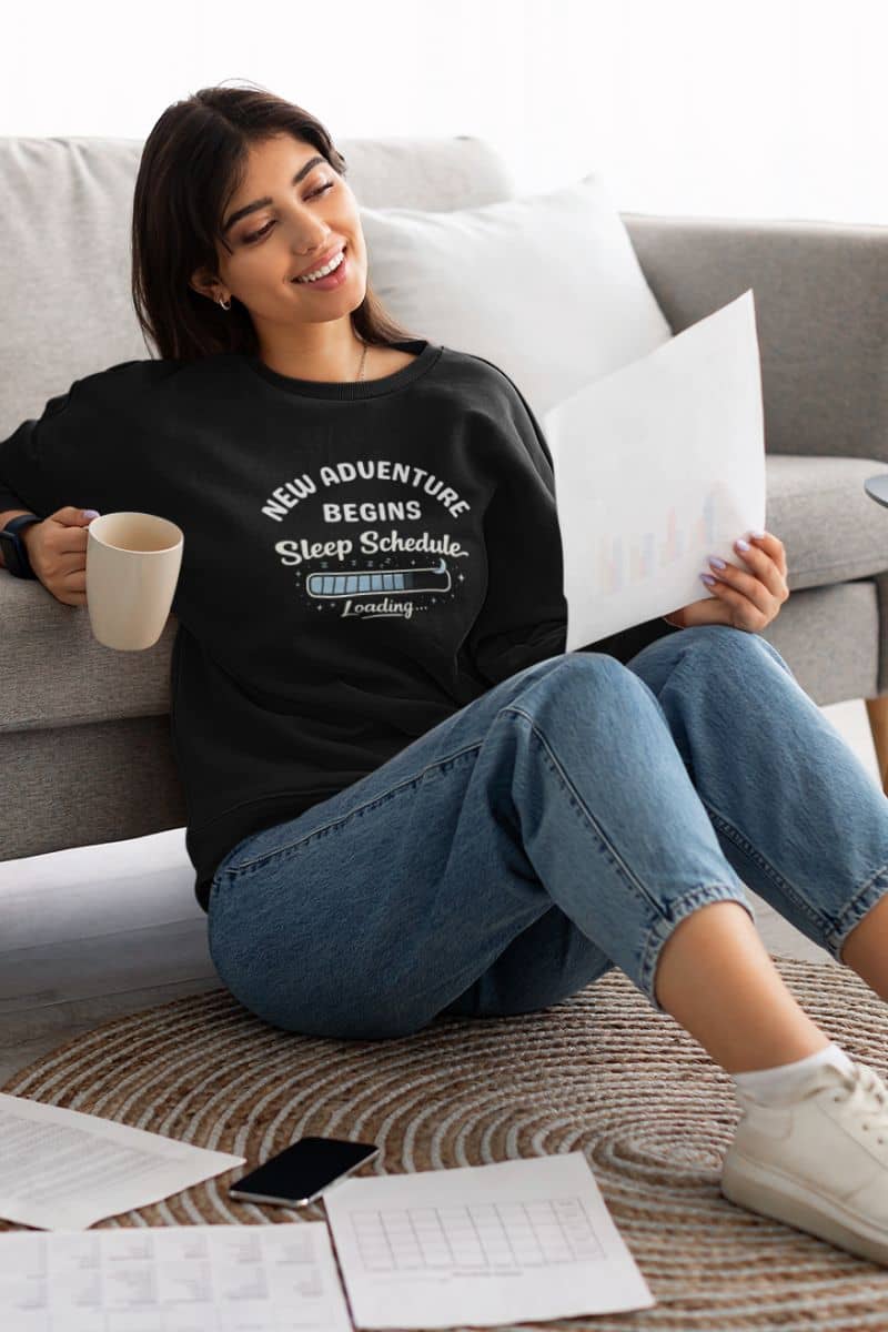 always tired pullover worn by a woman wearing a black sweatshirt with text 'New Adventure Begins, Sleep Schedule Loading…'. sitting on a couch holding a coffee cup and reading a document