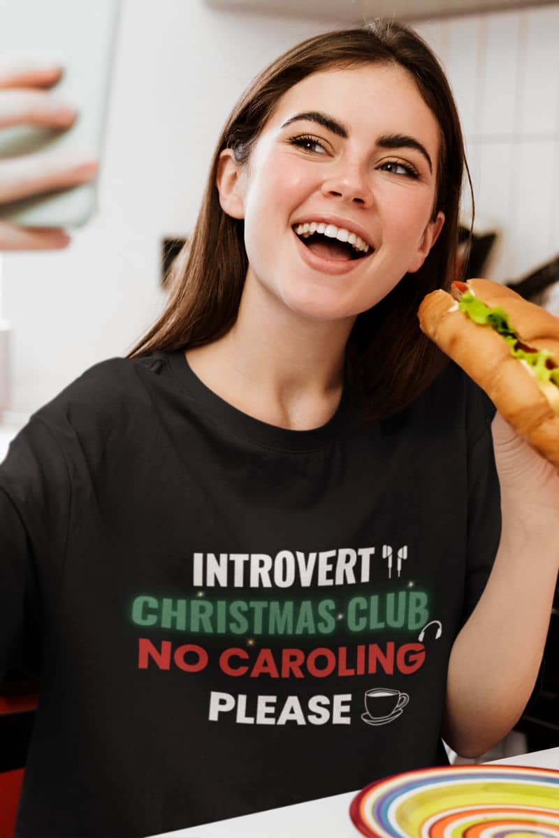 anti social shirt with a girl in a black t-shirt with text 'Introvert Christmas Club, No Caroling Please', taking a selfie while eating bread