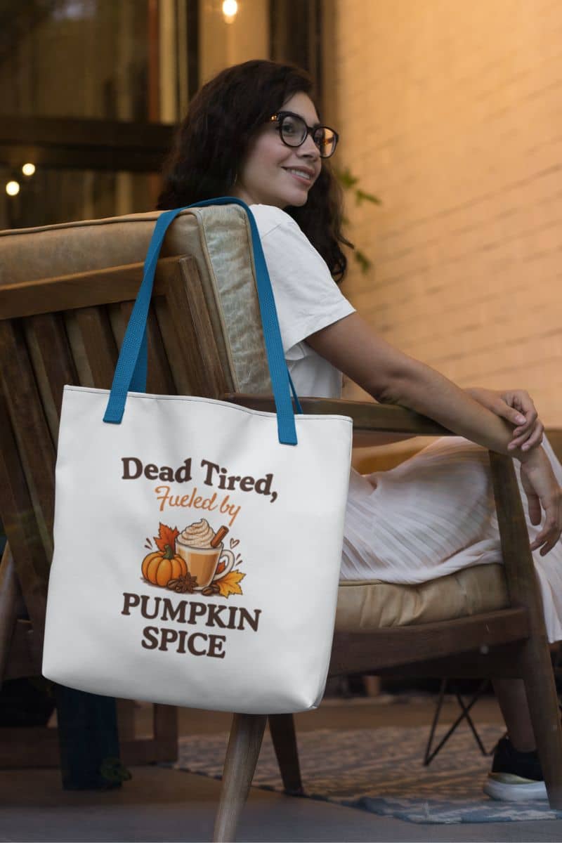 A autumn tote bag featuring pumpkins, autumn leaves, a hot drink, and the text 'Dead Tired, Fueled by Pumpkin Spice', next to a woman sitting on a wooden chair in a warm indoor setting.