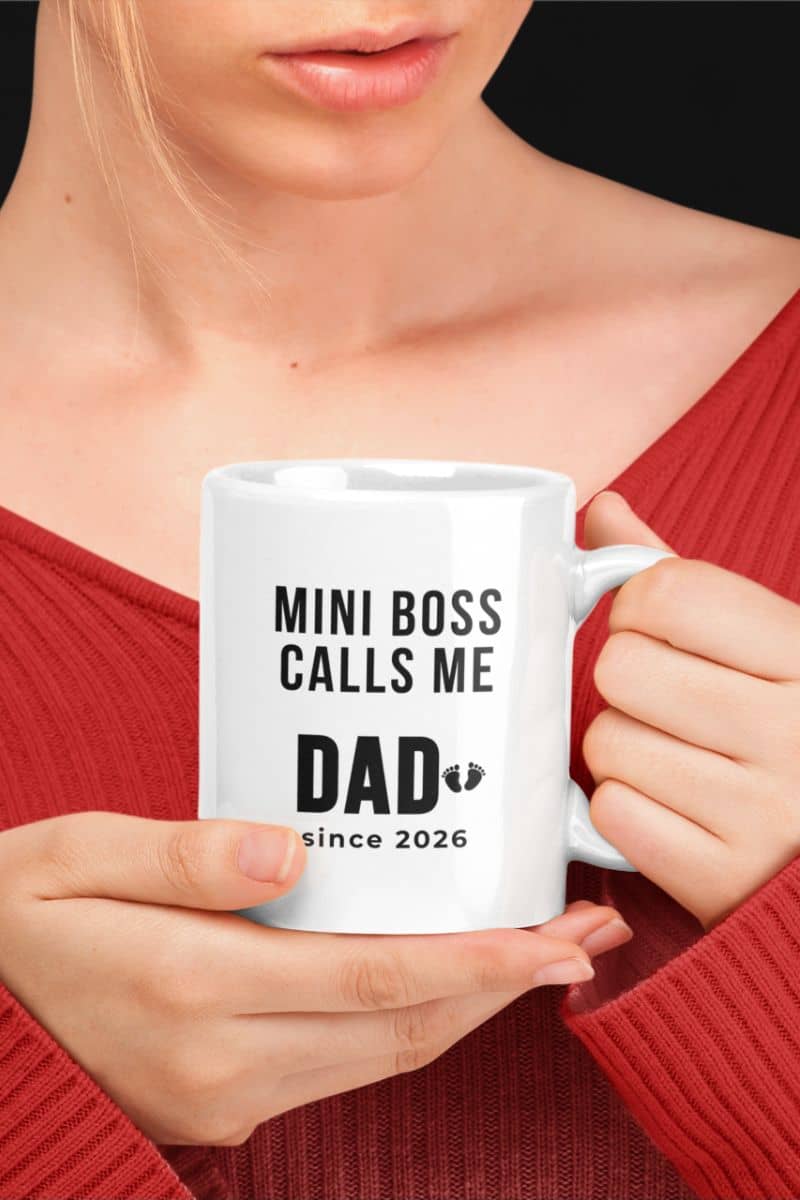 baby announcement mug with a person holding a white mug with text 'MINI BOSS CALLS ME DAD since 2026' against a neutral background