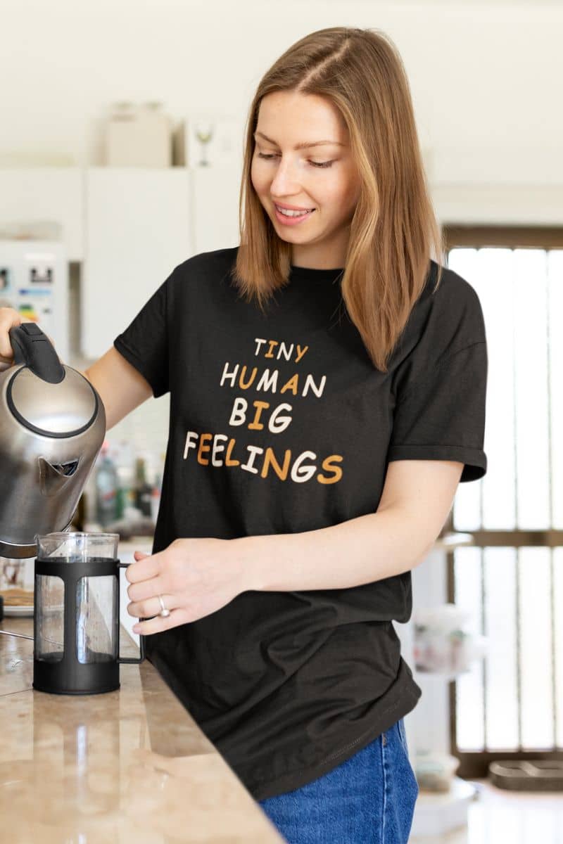 back to school shirt with a woman in a kitchen wearing a black graphic t-shirt with the text 'Tiny Human Big Feelings' in gold and white, pouring water from an electric kettle into a French press.