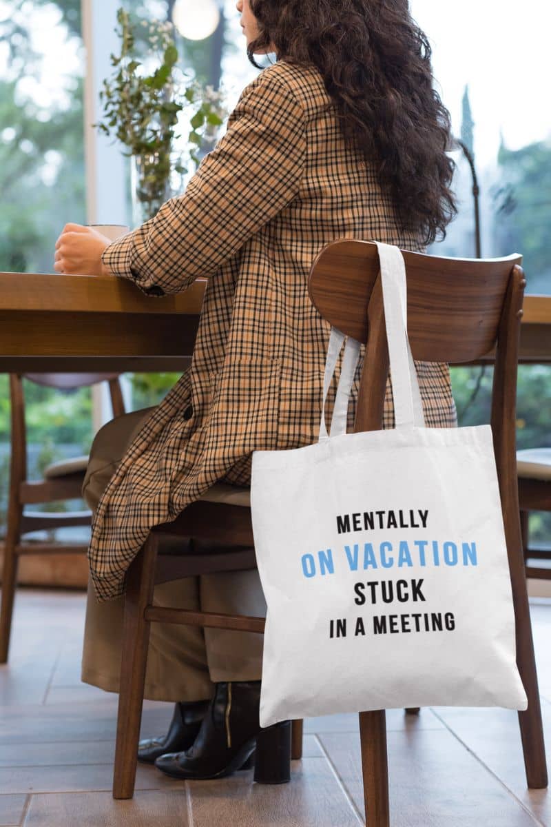 beach tote bag with a person sitting at a table with a tote bag that says 'Mentally on vacation, stuck in a meeting' in a casual setting.