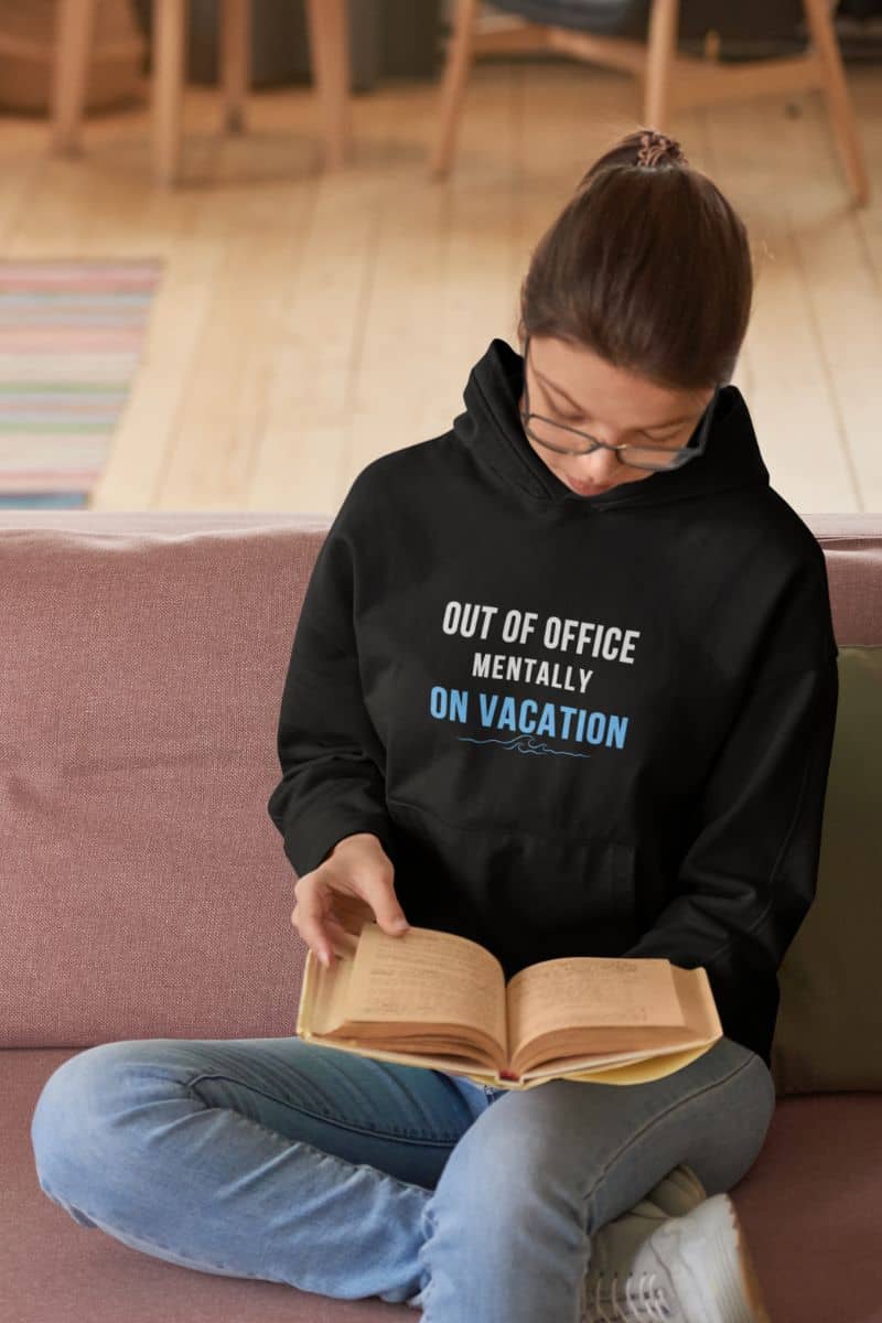 beach vacation hoodie with a person wearing a black hoodie with text 'Out of Office: Mentally on Vacation', sitting on a couch reading a book.