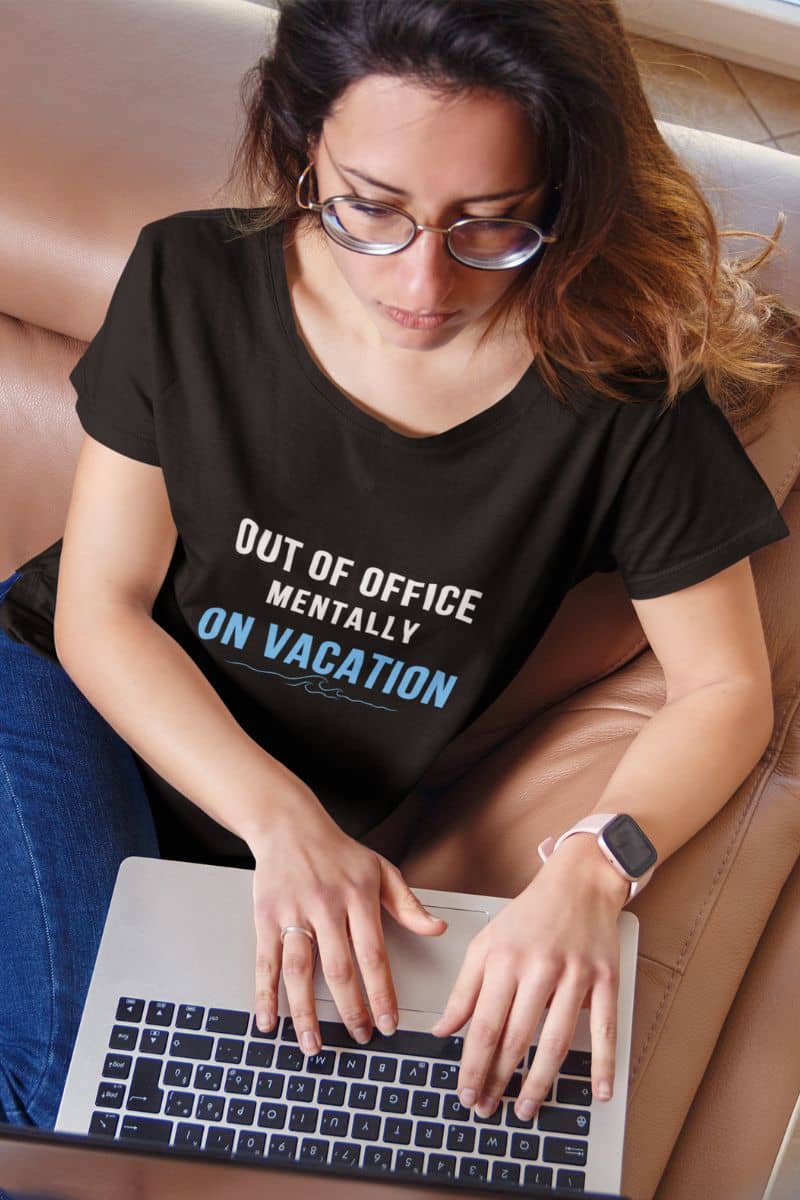 beach vacation tee with a woman wearing a black t-shirt with text 'Out of Office: Mentally on Vacation', using a laptop.