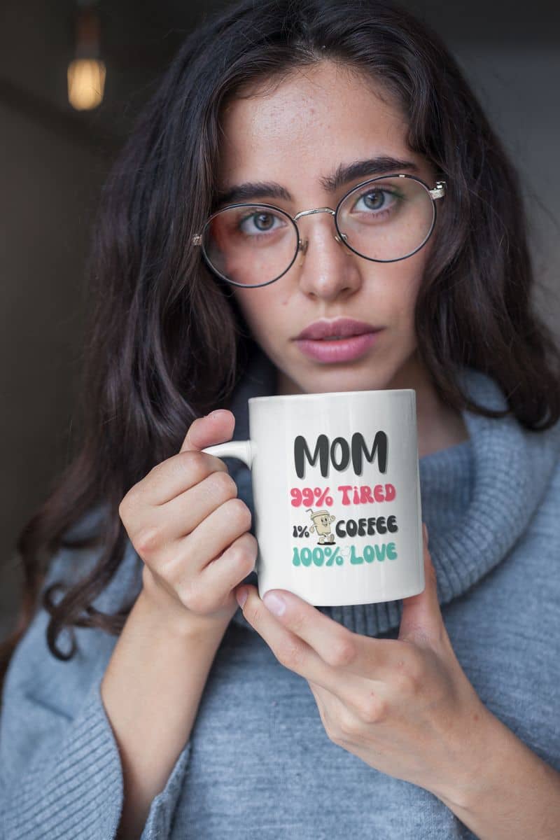 best mom coffee mug with a woman holding a mug with text 'Mom 99% Tired, 1% Coffee, 100% Love', wearing glasses and a gray sweater.
