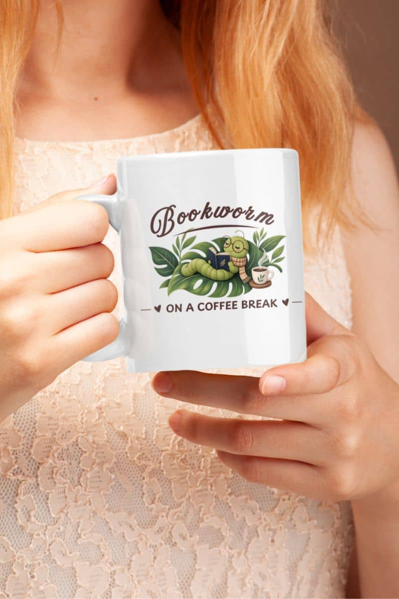 book lover mug with a person holding a white ceramic mug featuring the text 'Bookworm ON A COFFEE BREAK'. The person is wearing a light beige lace top, and the background is a soft, warm brown.