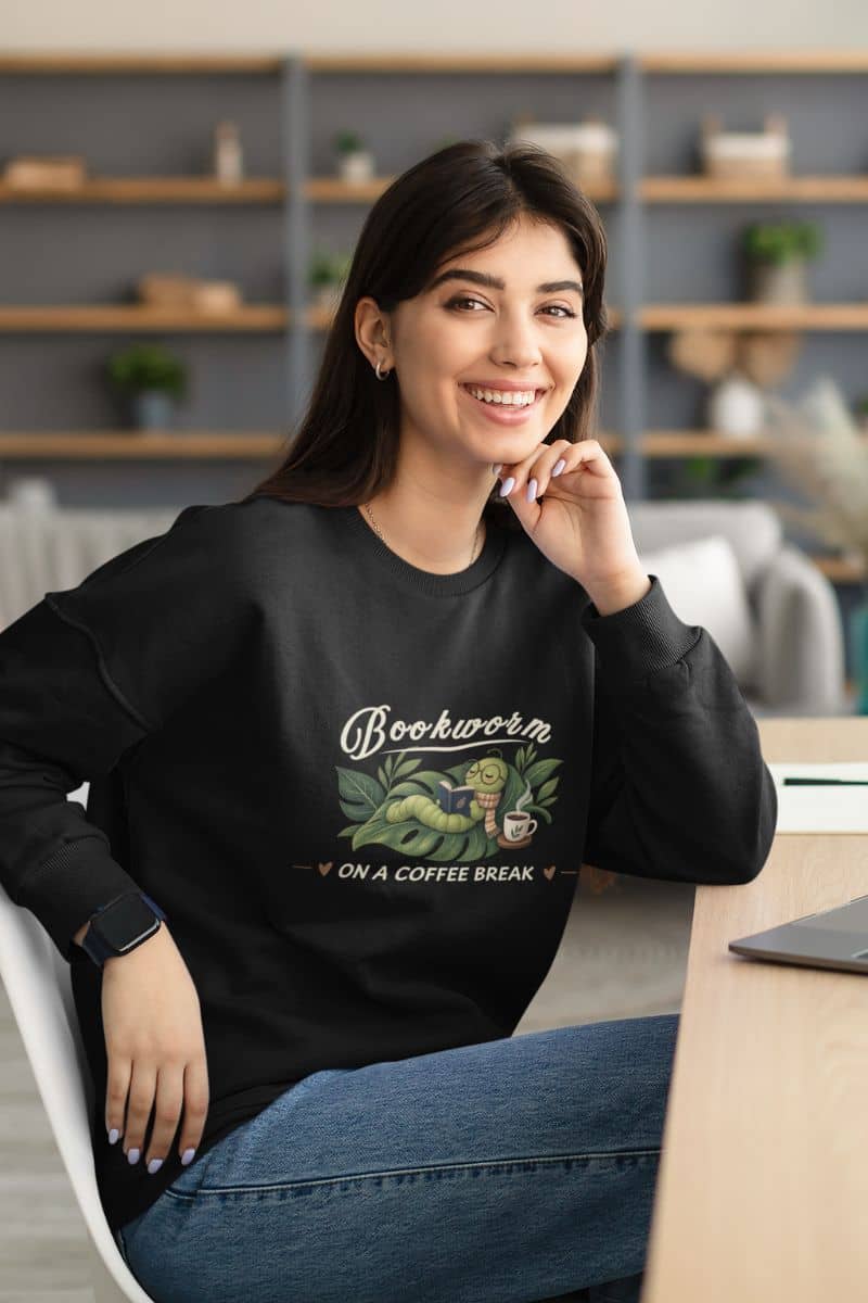 book sweatshirt with a woman sitting at a desk in a modern office setting, wearing a black crewneck sweatshirt  that reads 'Bookworm ON A COFFEE BREAK' with an illustration of a bookworm in a coffee cup.
