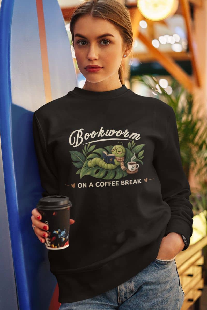 book themed sweatshirt with a woman wearing a black sweatshirt featuring the text 'Bookworm ON A COFFEE BREAK'. She is holding a takeaway coffee cup and wearing blue jeans, set against a warm, blurred indoor background.