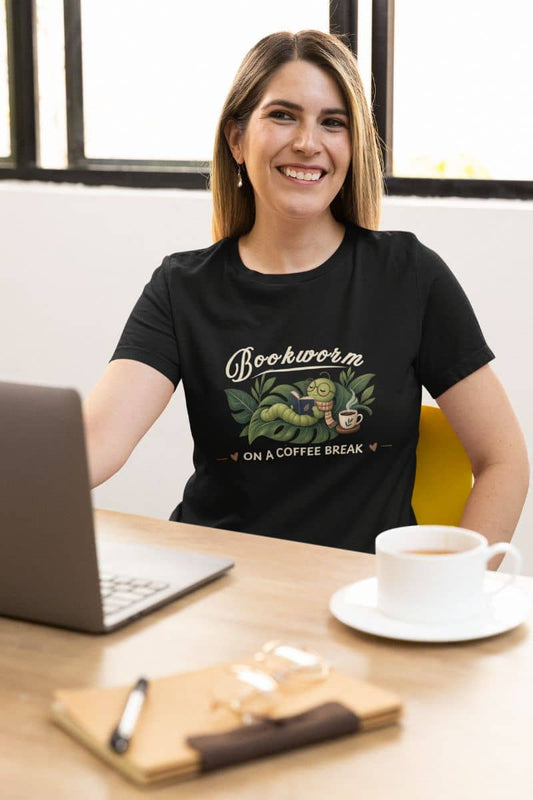 bookworm t shirt with woman wearing a black graphic T-shirt that reads 'Bookworm on a coffee break'. sitting at the desk has a laptop, a white coffee cup, and a notebook.