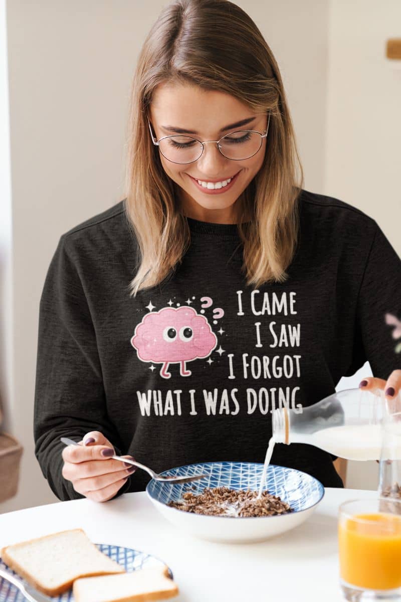 brain joke sweatshirt with a woman wearing a black sweatshirt with a pink brain graphic and text 'I came, I saw, I forgot what I was doing', eating cereal.