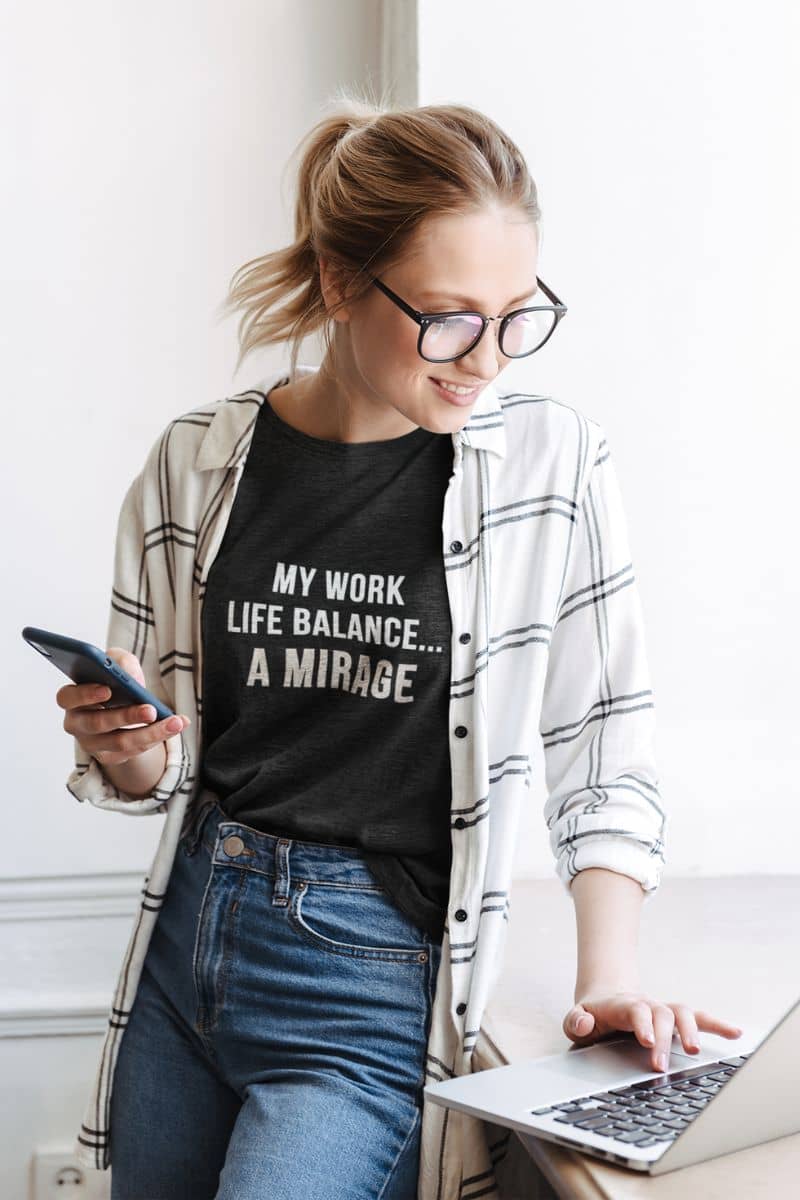 burnout humor shirt with a woman wearing a black t-shirt with text 'My Work-Life Balance Is a Mirage', using a laptop and smartphone indoors.